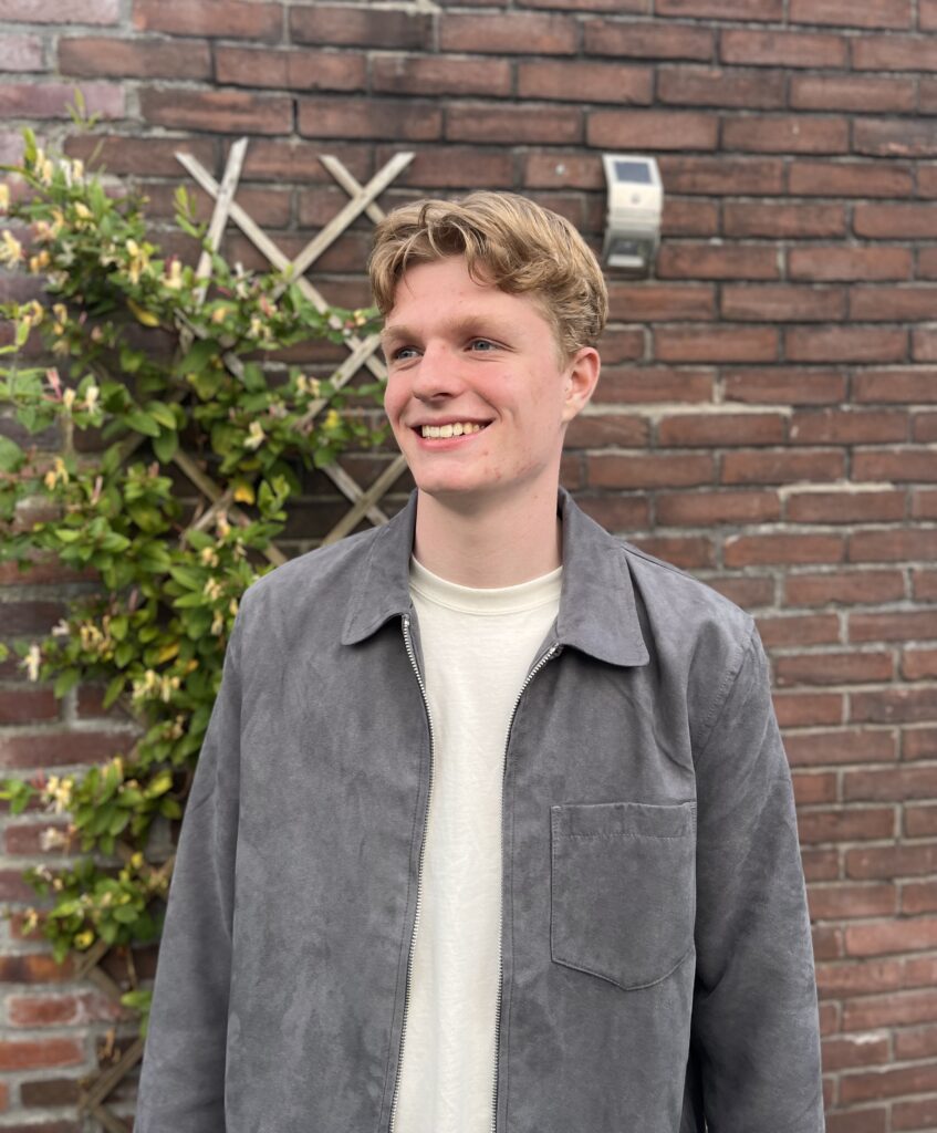 Portretfoto van een blonde, blanke jongen met een vriendelijke glimlach, staand in een tuin met groene planten op de achtergrond.